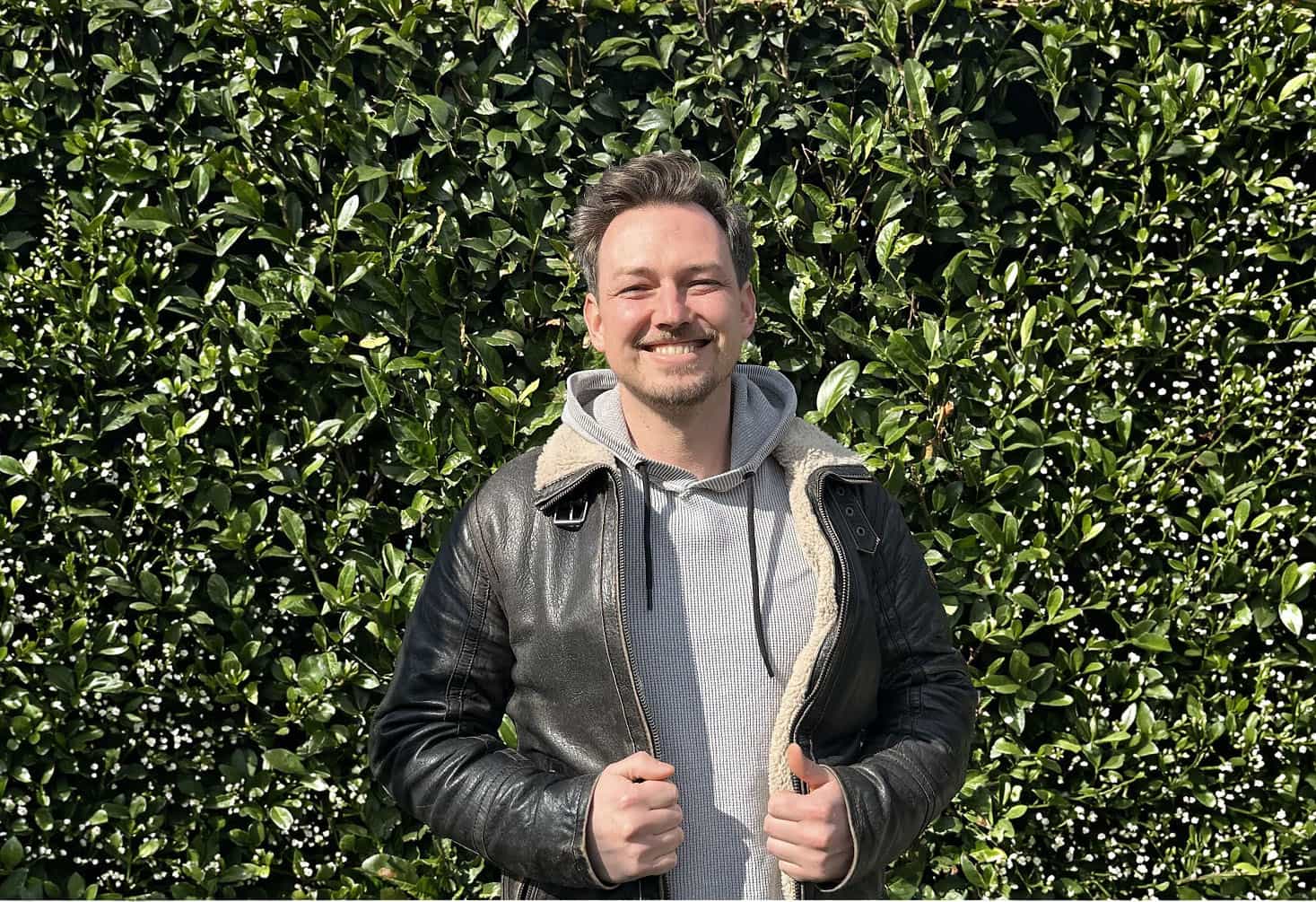 Lukas Blaik, Heilpraktiker für Psychotherapie und Coach, lächelt in Lederjacke und Kapuzenpullover vor einer grünen Hecke