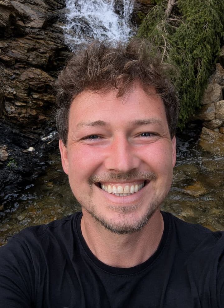 Lukas Blaik lächelt breit in die Kamera, Selfie vor einem kleinen Wasserfall und Felsen im Wald